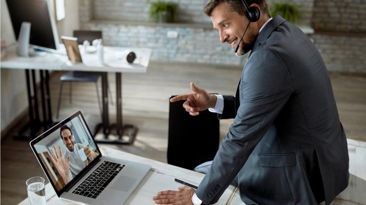 A man in a suit wearing a headset points at his laptop screen during a video call in a modern office