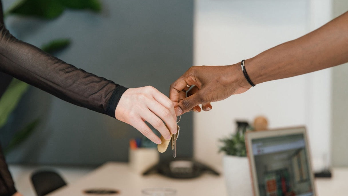 Two people exchanging a set of keys across an office desk with a laptop in the background.