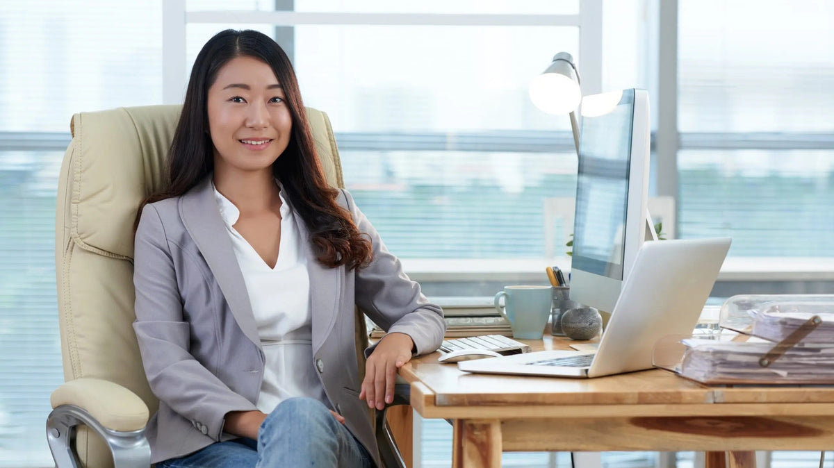Woman in a blazer seated at an office desk beside large windows, with a monitor and laptop