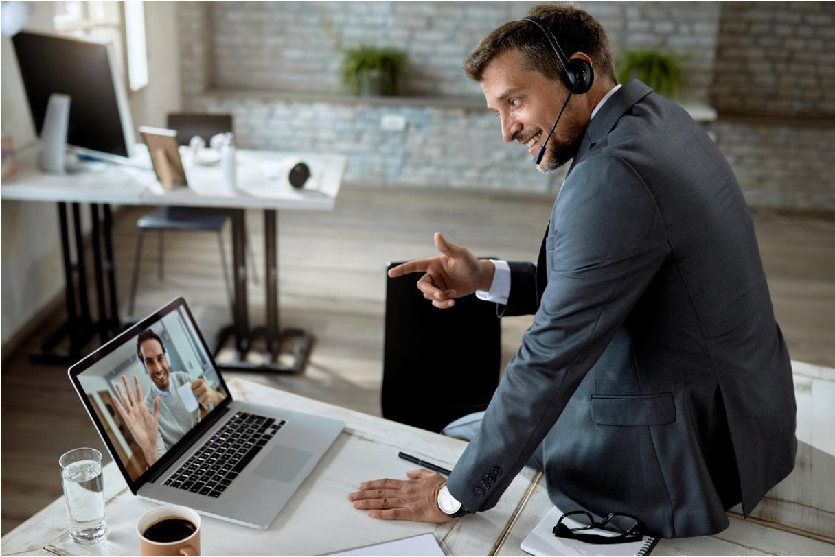 A man in a suit wearing a headset points at his laptop screen during a video call in a modern office
