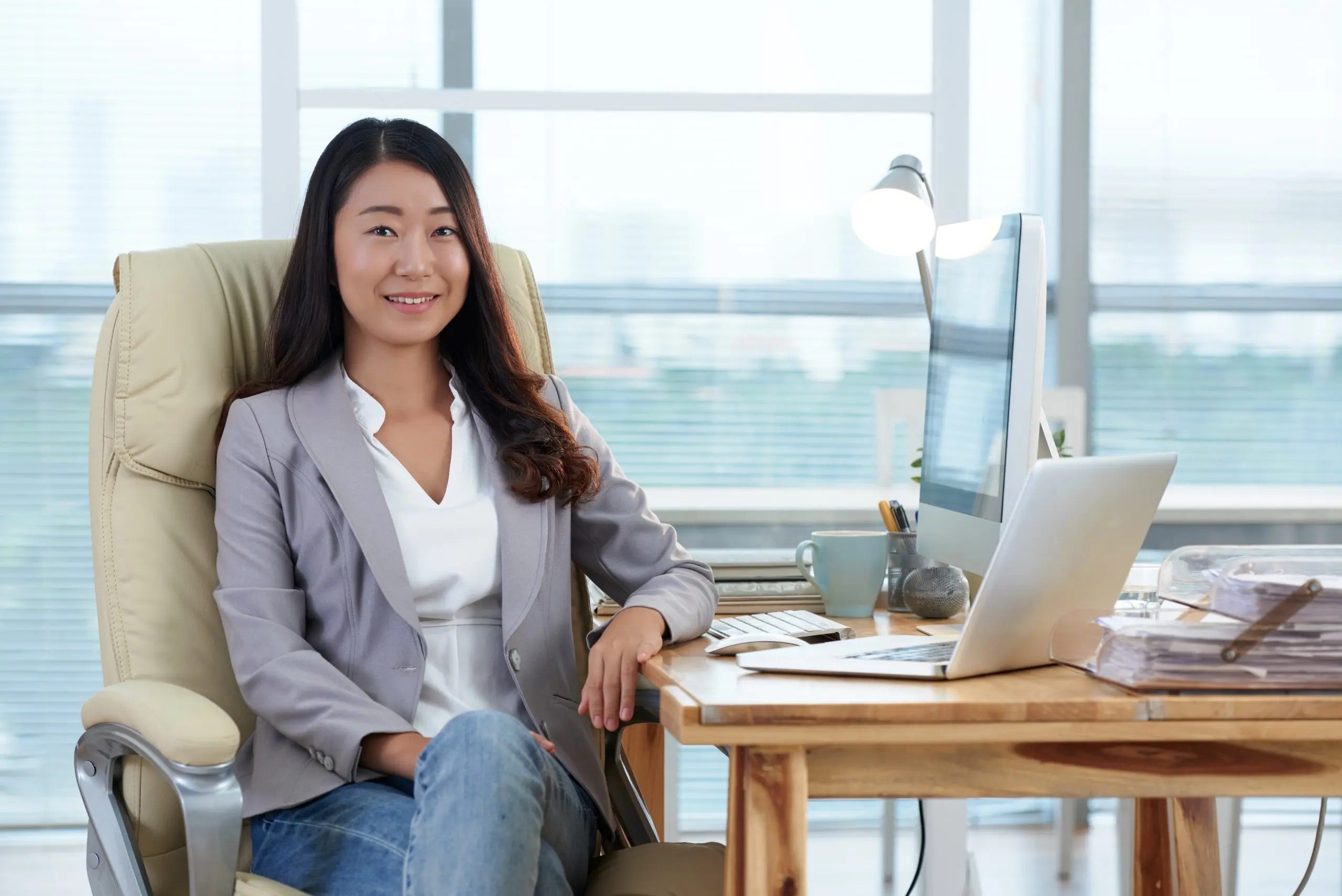 Woman in a blazer seated at an office desk beside large windows, with a monitor and laptop